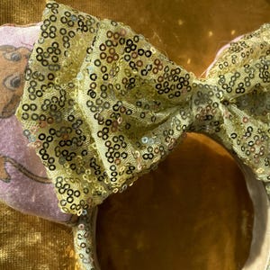 May include: A pair of mouse ear headbands. One ear is light purple with a cartoon illustration. The other ear is covered in gold sequins. A large bow made of gold sequins sits between the ears. The headband is white.