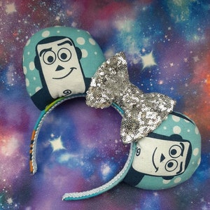 May include: A pair of blue and white mouse ears with a silver sequin bow. The ears feature a cartoon character with a white face and black eyes.