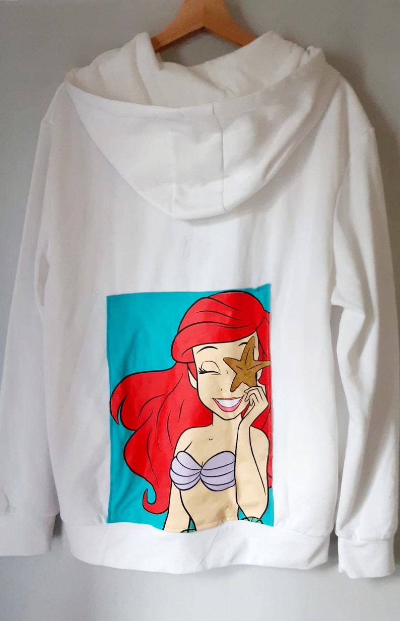 Puede incluir: Sudadera con capucha blanca con un gr&aacute;fico de dibujos animados de una sirena en la espalda. La sirena tiene el pelo rojo y sostiene una estrella de mar. El gr&aacute;fico est&aacute; sobre un fondo turquesa. La sudadera tiene capucha con cord&oacute;n y mangas largas.