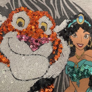 Op de afbeelding: Grijs T-shirt met een pailletten illustratie van Rajah de tijger en prinses Jasmine uit Aladdin. Rajah is oranje, wit en roze. Jasmine draagt een turquoise outfit en gouden sieraden. De achtergrond heeft een zebrapatroon.