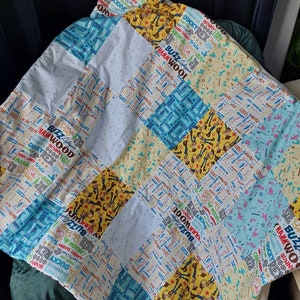 Puede incluir: Una colcha de retazos en colores azul, blanco y amarillo. La colcha est&aacute; hecha de diferentes telas con un patr&oacute;n repetido de las palabras "Buzz", "Woody", "Rex" y "Ducky".