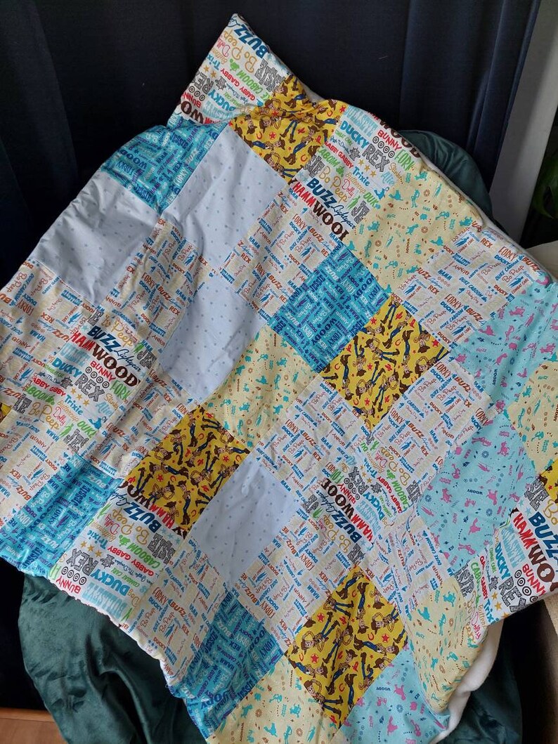 Puede incluir: Una colcha de retazos en colores azul, blanco y amarillo. La colcha est&aacute; hecha de diferentes telas con un patr&oacute;n repetido de las palabras "Buzz", "Woody", "Rex" y "Ducky".