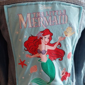 Op de afbeelding: Een lichtblauw denim jack met een blauwe en witte patch met Ariel, de Kleine Zeemeermin, uit de Disney-film. De patch heeft de tekst "Disney De Kleine Zeemeermin" erop.