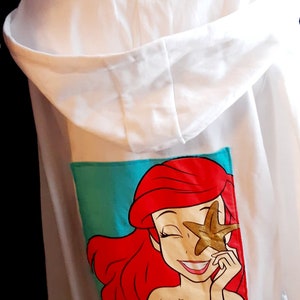 Puede incluir: Sudadera con capucha blanca con un gr&aacute;fico colorido de una sirena de dibujos animados con cabello rojo y una estrella de mar dorada. El gr&aacute;fico est&aacute; sobre un fondo turquesa. La sudadera tiene capucha con cord&oacute;n y mangas largas.