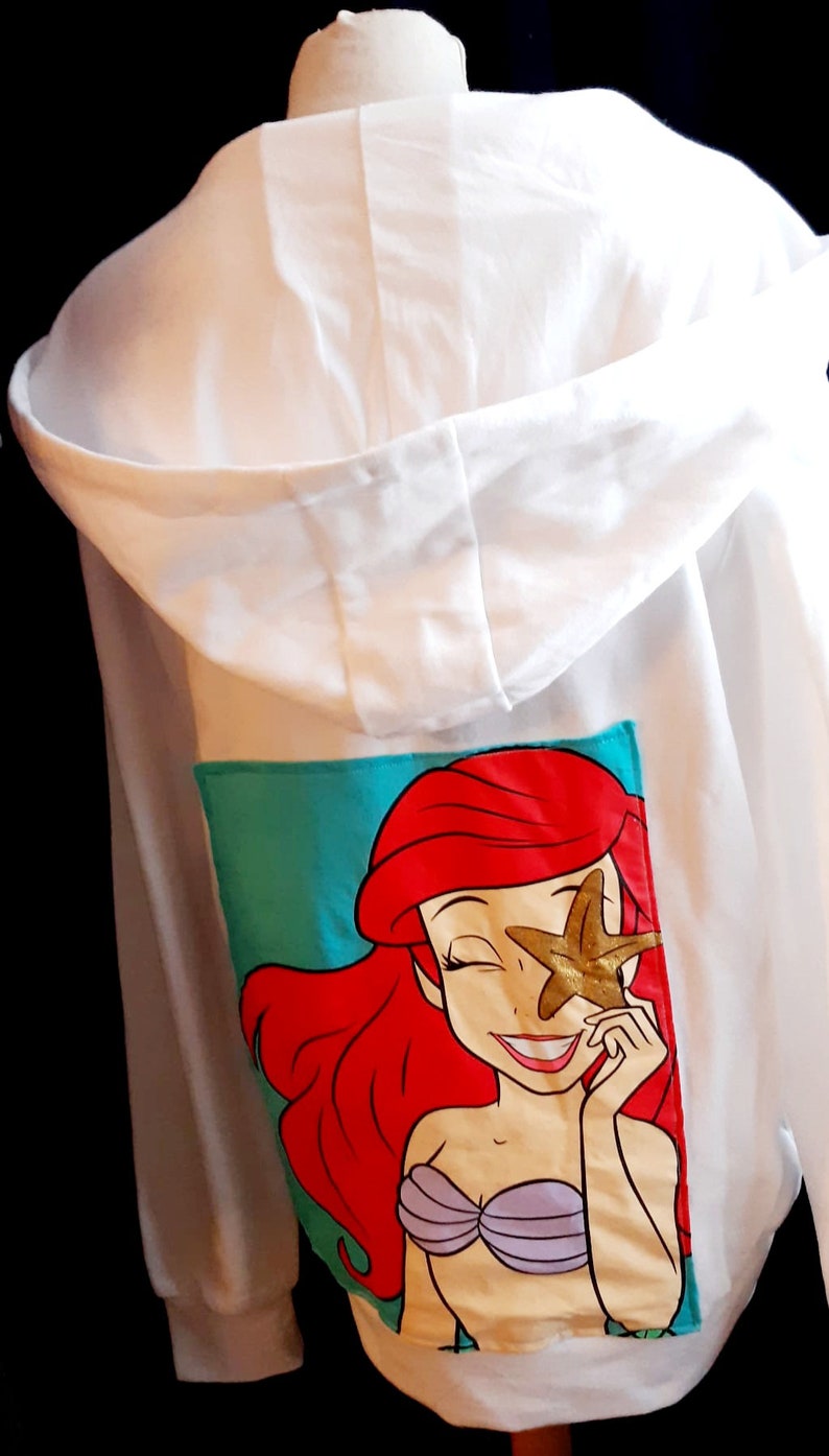 Puede incluir: Sudadera con capucha blanca con un gr&aacute;fico colorido de una sirena de dibujos animados con cabello rojo y una estrella de mar dorada. El gr&aacute;fico est&aacute; sobre un fondo turquesa. La sudadera tiene capucha con cord&oacute;n y mangas largas.