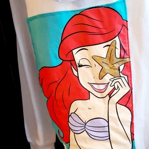 Puede incluir: Sudadera con capucha blanca con un bolsillo turquesa con una ilustraci&oacute;n de dibujos animados de Ariel de La Sirenita. Ariel tiene el pelo rojo, un sujetador de concha p&uacute;rpura y sostiene una estrella de mar dorada sobre un ojo. La sudadera tiene una capucha con cord&oacute;n.