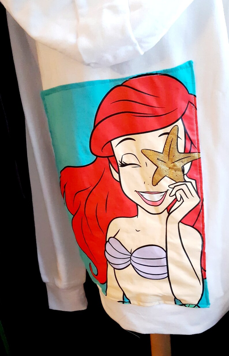 Puede incluir: Sudadera con capucha blanca con un bolsillo turquesa con una ilustraci&oacute;n de dibujos animados de Ariel de La Sirenita. Ariel tiene el pelo rojo, un sujetador de concha p&uacute;rpura y sostiene una estrella de mar dorada sobre un ojo. La sudadera tiene una capucha con cord&oacute;n.