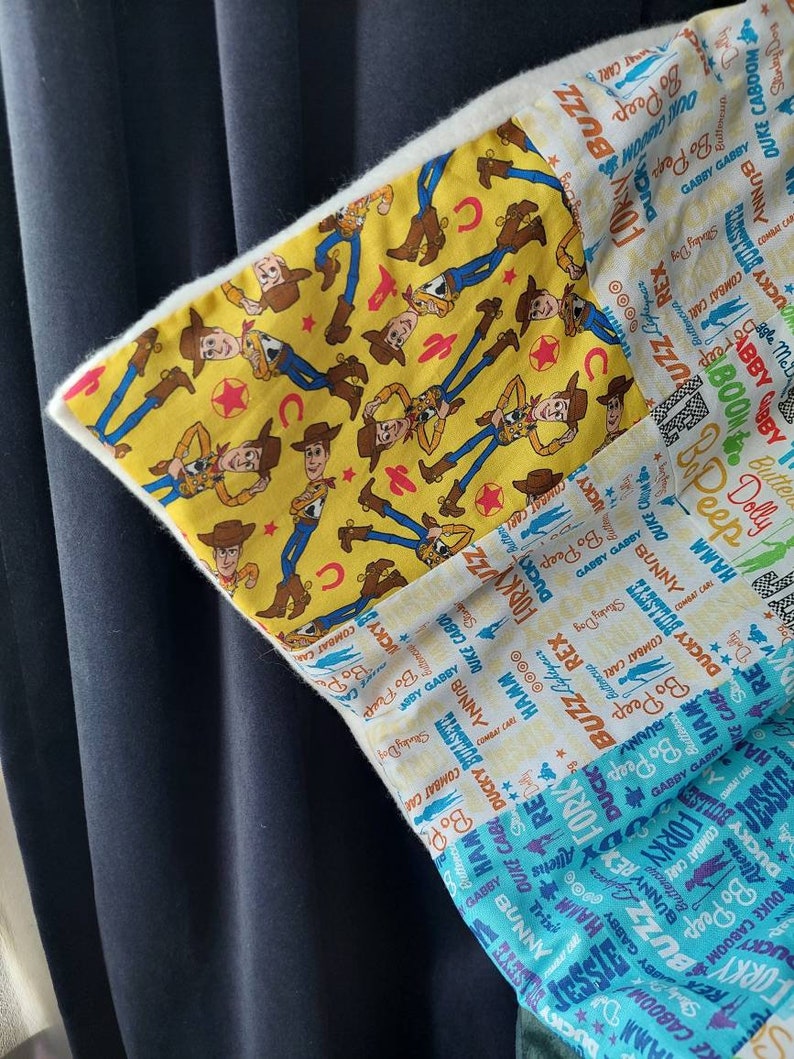 Puede incluir: Una almohada de tela blanca con un dise&ntilde;o de patchwork que presenta una tela amarilla con un patr&oacute;n repetido de Woody y Buzz Lightyear, personajes de la franquicia Toy Story. Los otros paneles de tela presentan un patr&oacute;n azul y blanco con los nombres de los personajes de la franquicia Toy Story.