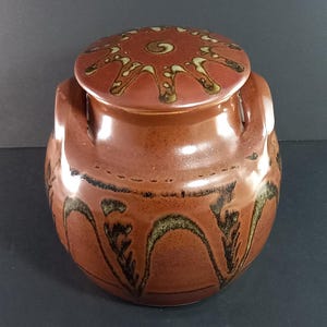 May include: A brown ceramic jar with two handles and a lid. The jar features a decorative design of brown and green swirls. The lid has a red and brown swirl design.