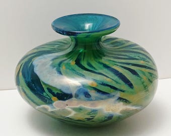 May include: A squat, rounded glass vase featuring a swirling pattern of deep blue, emerald green, and white streaks. The vessel has a wide, flat-rimmed opening and a smooth, polished surface with small yellow speckles embedded throughout the glass.