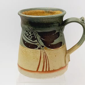 May include: Handmade ceramic mug with a unique design. The mug features a green and brown floral pattern on a cream-coloured base. The handle and rim are also green, with a touch of orange at the base. A rustic, artistic piece.