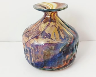 May include: A decorative glass vase with a rounded body and a flared rim. The vase has a swirling pattern of colours, including shades of blue, purple, gold, and brown. The glass has a textured appearance, adding visual interest to the piece.