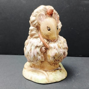 May include: A ceramic figurine of a mouse with light brown fur, wearing a textured, ruffled white and tan garment. The mouse is depicted in a standing position on a rounded base, with detailed facial features and a small orange accent on its head.