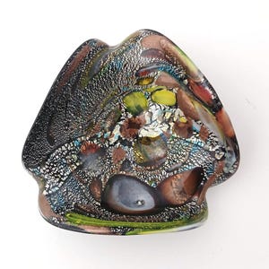 May include: A decorative glass dish with an unusual, organic form. The dish has a mix of colours, including black, brown, green, and yellow, with a speckled, textured look. The design has a mix of opaque and translucent elements.