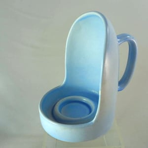May include: A light blue ceramic candle holder with a handle. The holder has a curved back and a circular base to hold a candle. The handle is on the right side of the holder. The candle holder is a light blue colour.
