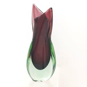 May include: A decorative glass vase with a distinctive shape and colour scheme. The vase features a deep burgundy centre, transitioning to green at the base, with a flared opening. The glass appears to be hand-blown.