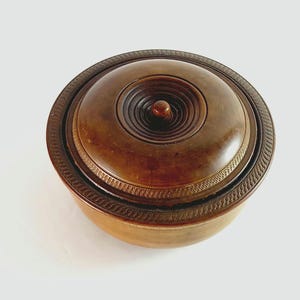 May include: A round wooden bowl with a lid. The bowl is dark brown and has a carved design around the rim. The lid has a knob in the centre and a concentric circle design.