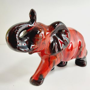 May include: A ceramic elephant ornament with a glossy finish. The elephant is mainly red with dark brown and black accents. The trunk is raised, and the ears are large. A decorative item.