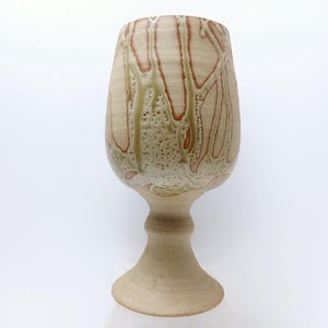 May include: A beige ceramic goblet with a wide bowl and a pedestal base. The goblet has a textured surface with brown and green drip-like patterns. The overall design is organic and handcrafted, suitable for display or use.