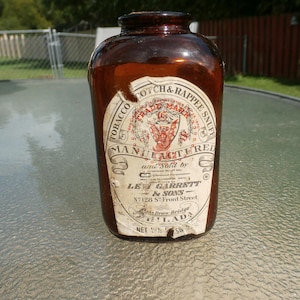 Puede incluir: Una botella de vidrio marrón con una etiqueta blanca que dice "TOBACCO SCOTCH & RAPPEE SNUFF MANUFACTURED and sold by LEVI GARRETT & SONS N128 ST Front Street  Goulds Draw Bridge PHILADELPHIA NET 1/4 5/8"