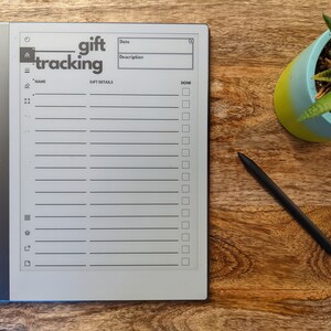 K&ouml;nnte beinhalten: Ein grauer E-Reader mit einem wei&szlig;en Bildschirm, auf dem eine Geschenk-Tracking-Liste angezeigt wird. Die Liste enth&auml;lt Felder f&uuml;r Name, Datum, Beschreibung und ein Kontrollk&auml;stchen f&uuml;r erledigt. Der E-Reader befindet sich auf einer Holzoberfl&auml;che mit einem schwarzen Stift und einer Topfpflanze.