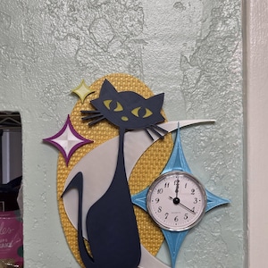 Könnte beinhalten: Retro-Wanduhr mit einer marineblauen Katzensilhouette und gelben Augen, vor einem goldfarbenen, strukturierten Hintergrund. Die Uhr hat ein weißes Zifferblatt mit schwarzen Zahlen und Zeigern sowie einen blauen Sternrahmen. Misst ca. 25 cm.