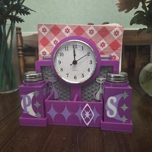 Puede incluir: Un juego de reloj y salero y pimentero morado. El reloj tiene una esfera blanca con números y manecillas negras. Los saleros y pimenteros están etiquetados con las letras "P" y "S". El juego está decorado con formas geométricas y siluetas de gatos.
