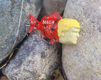 May include: A red and black fishing lure shaped like a fish with a human face. The lure has the word "MAGA" written on it.