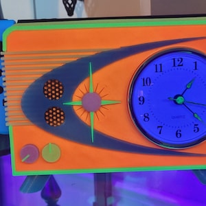 May include: A retro-style clock radio with a bright orange body, green trim, and a blue clock face. The clock face has black numbers and hands. The radio has two knobs on the front.