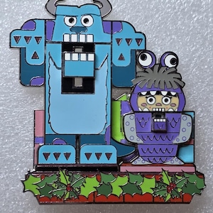 May include: A decorative pin featuring two cartoon characters, one tall and blue with horns, the other small and purple. The pin is set on a red brick base with green holly and red berries.