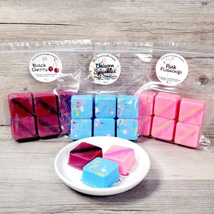 May include: Three bags of wax melts, each with six square wax melts. The bags are labeled "Black Cherry", "Unicorn Sprinkles", and "Pink Flamingo". There are three wax melts on a white plate, one of each scent.