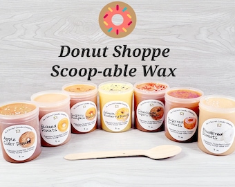 Könnte beinhalten: Sechs verschiedene Duftwachs-Schmelzen in durchsichtigen Behältern mit Etiketten, auf denen "Apple Cider Donut", "Glazed Donuts", "Sugary Pumpkin Donuts", "Lemon Blueberry Donuts", "Cinnamon Sugar Donuts" und "Powdered Donuts" steht. Die Wachs-Schmelzen sind in einer Reihe auf einer Holzoberfläche angeordnet. Ein Holzlöffel befindet sich vor den Behältern.