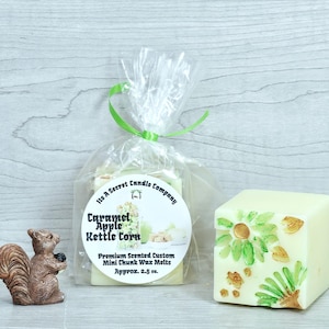 May include: A brown squirrel figurine stands next to a clear plastic bag containing Caramel Apple Kettle Corn wax melts, tied with a green ribbon. A single square, off-white wax melt with green and gold leaf-like accents sits to the right.