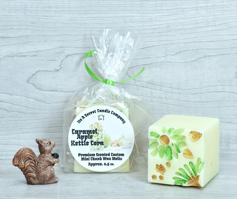 May include: A brown squirrel figurine stands next to a clear plastic bag containing Caramel Apple Kettle Corn wax melts, tied with a green ribbon. A single square, off-white wax melt with green and gold leaf-like accents sits to the right.