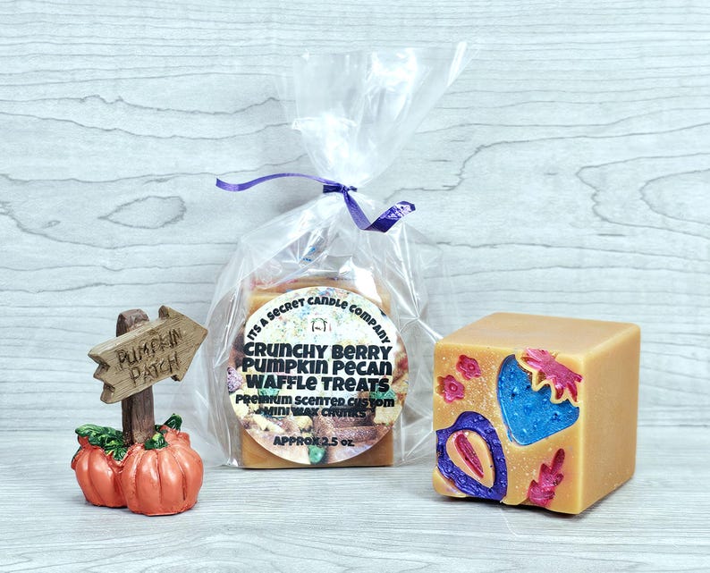 May include: A small wooden pumpkin patch sign, a clear plastic bag containing wax chunks with a label reading Its A Secret Candle Company Crunchy Berry Pumpkin Pecan Waffle Treats, and a tan wax cube with blue and purple berry-shaped embeds.
