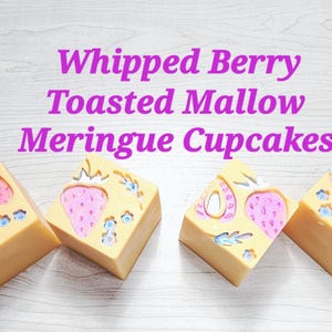 May include: Four square-shaped bars of soap in a light tan colour, each featuring embossed pink strawberry designs with blue accents. The text "Whipped Berry Toasted Mallow Meringue Cupcakes" is printed in purple letters above the soap bars.