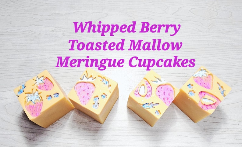 May include: Four square-shaped bars of soap in a light tan colour, each featuring embossed pink strawberry designs with blue accents. The text "Whipped Berry Toasted Mallow Meringue Cupcakes" is printed in purple letters above the soap bars.