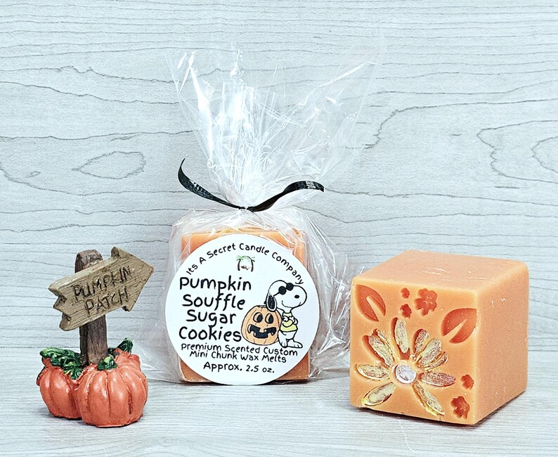 May include: A clear plastic bag containing orange wax melts with a label reading Its A Secret Candle Company Pumpkin Souffle Sugar Cookies, next to a small pumpkin patch sign and a single orange wax cube with a floral design.