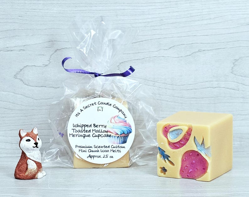 May include: A small fox figurine next to a clear plastic bag containing wax melts with a label reading Its A Secret Candle Company Whipped Berry Toasted Mallow Meringue Cupcake Premium Scented Custom Mini Chunk Wax Melts Approx 25 oz and a cube.