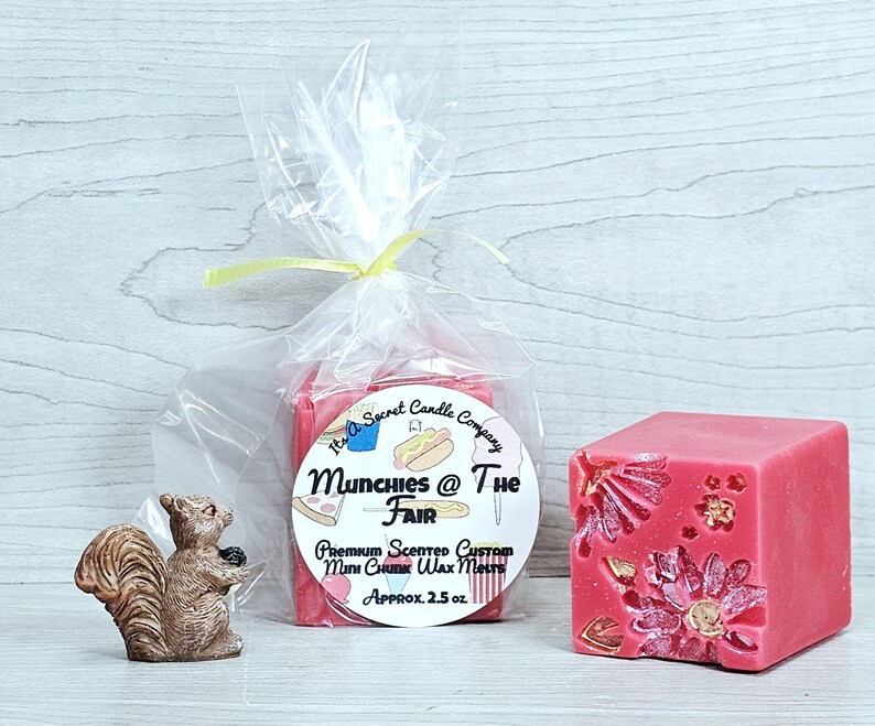 May include: A clear plastic bag containing pink wax melts with a label reading Munchies @ The Fair, Premium Scented Custom Mini Chunk Wax Melts, Approx. 71g. Next to it is a pink wax cube with floral indentations and a small brown squirrel figurine.