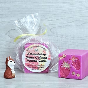 May include: A pink wax melt cube with embossed strawberry designs and gold accents, alongside a clear plastic bag containing more wax melts with a label reading Strawberry Pina Colada Pound Cake, and a small brown and white fox figurine.