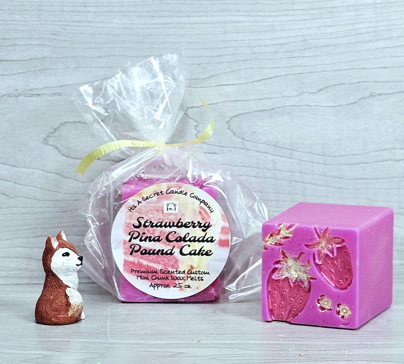 May include: A pink wax melt cube with embossed strawberry designs and gold accents, alongside a clear plastic bag containing more wax melts with a label reading Strawberry Pina Colada Pound Cake, and a small brown and white fox figurine.