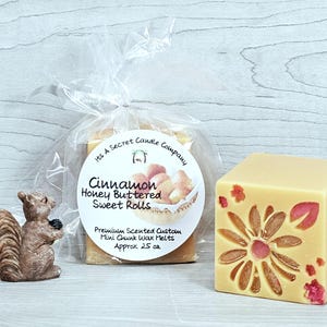 May include: A small brown squirrel figurine next to a clear plastic bag containing Cinnamon Honey Buttered Sweet Rolls wax melts with a round label, and a single beige wax melt cube featuring a pressed flower design with pink and red petals.