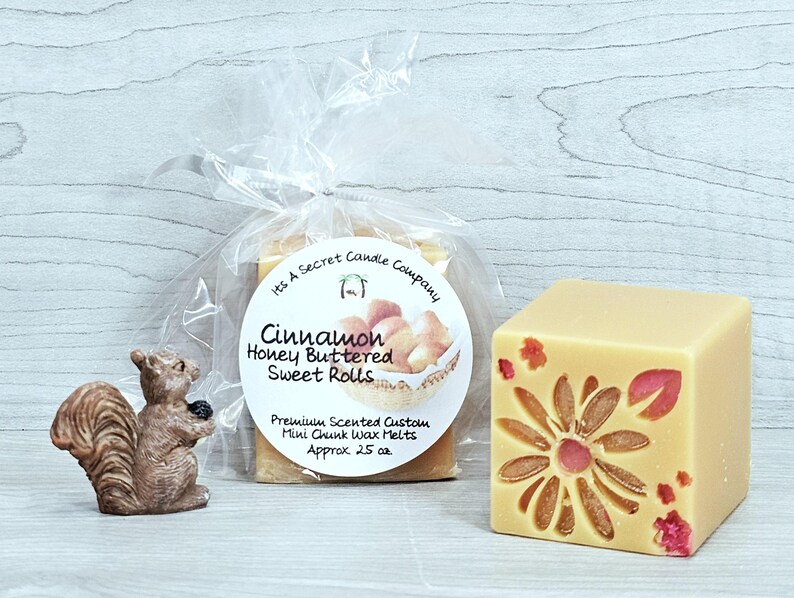 May include: A small brown squirrel figurine next to a clear plastic bag containing Cinnamon Honey Buttered Sweet Rolls wax melts with a round label, and a single beige wax melt cube featuring a pressed flower design with pink and red petals.