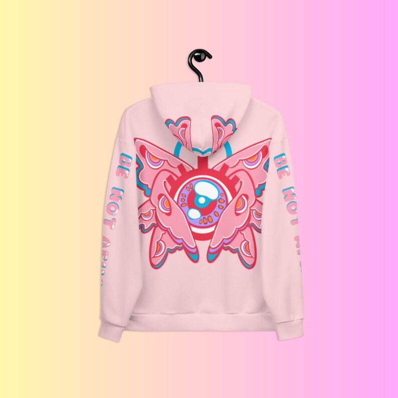 May include: A light pink hoodie with a large, colorful butterfly-eye design on the back. The design features shades of pink, blue, and red. The words "BE NOT AFRAID" are printed vertically on the left sleeve.