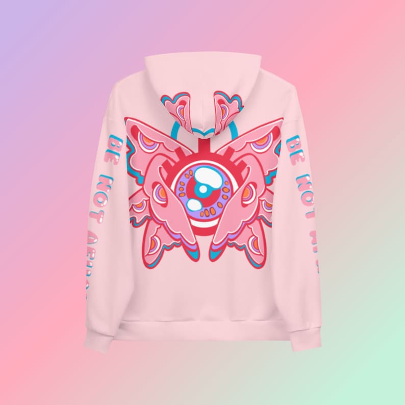 May include: A light pink hoodie with a large, colorful butterfly-eye design on the back. The design features pink, blue, and orange elements. The sleeves have the text "BE NOT AFRAID" printed vertically in blue.