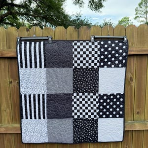May include: A black and white patchwork quilt with various patterns, including stripes, polka dots, and checkerboard designs. The quilt is hanging on a wooden fence. The quilt is approximately 102 cm wide and 127 cm long.
