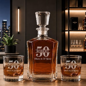 May include: A crystal whiskey decanter and two matching glasses. The decanter and glasses are engraved with the name "ROBERT" and the number "50", with the text "Cheers to 50 Years". The decanter is filled with amber liquid.