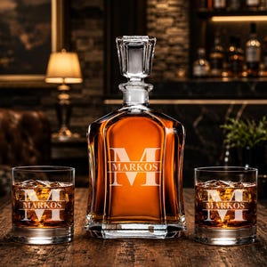 May include: A personalized whiskey decanter set with two matching glasses. The decanter and glasses are engraved with the name "MARKOS" and the letter "M". The set is filled with amber-colored liquid and sits on a wooden surface.