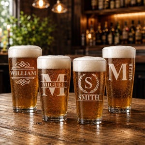 May include: Four clear pint glasses filled with beer and topped with white foam, each etched with a different name: William, Miguel, Smith, and Miller. The glasses sit on a wooden table in a bar setting, illuminated by overhead lights.